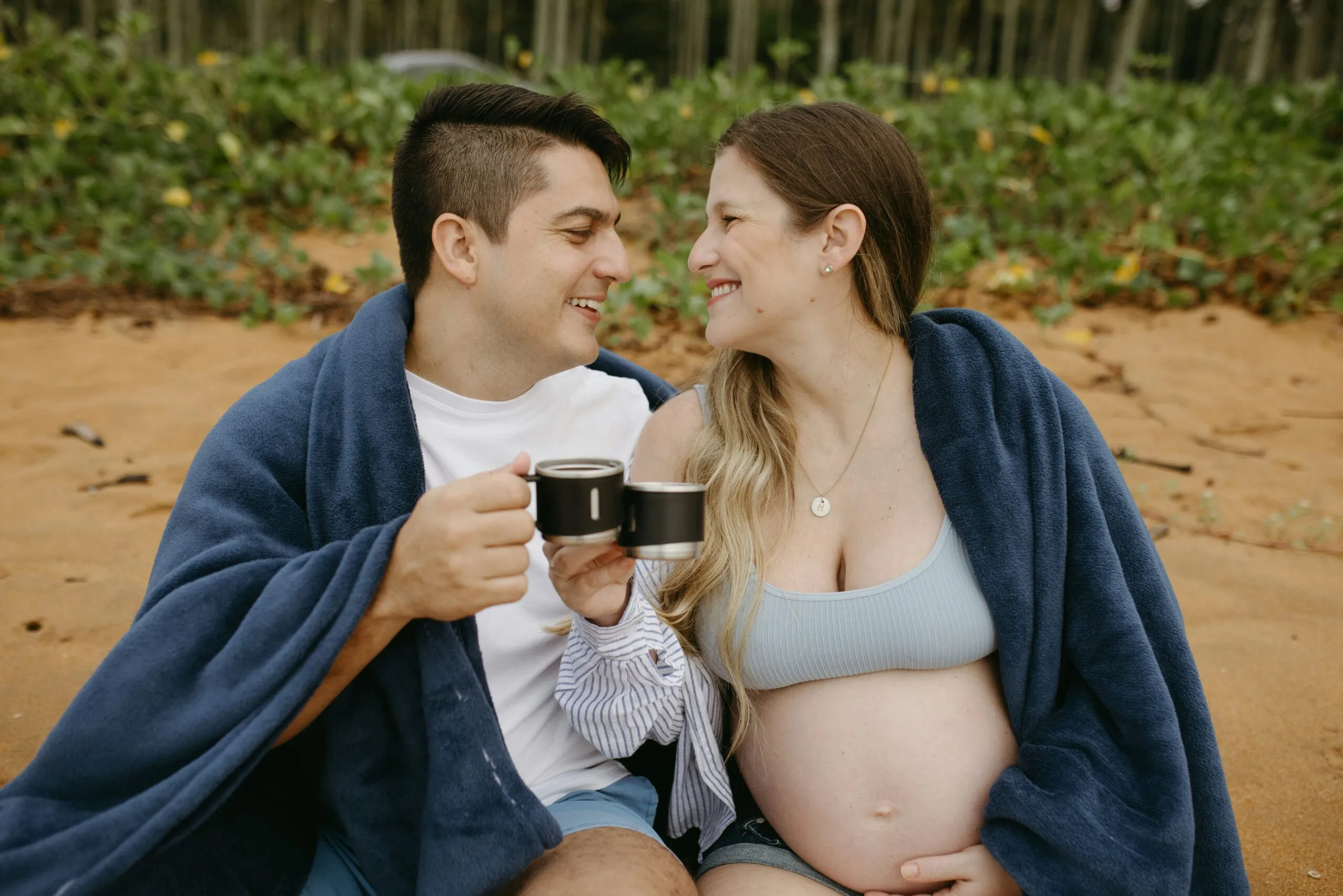 A pregnant couple sitting on a blanket holding a coffee cup