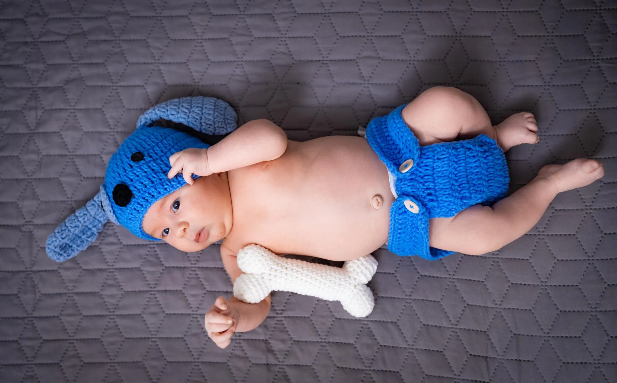 Baby dressed up in dog costume with plush bone toy A cute baby is lying on a quilt wearing a blue dog costume and holding a soft bone toy radiating joy