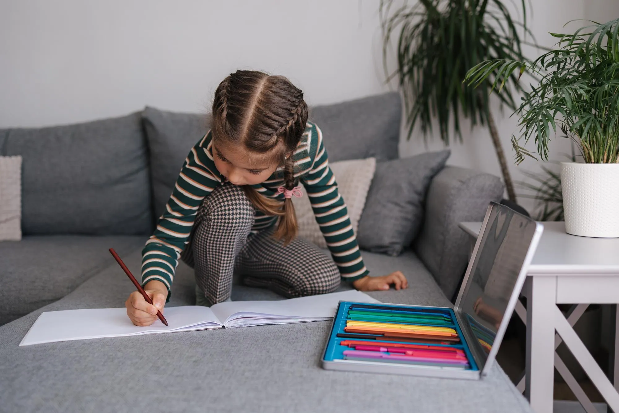Beautiful little girl is drawing in an album while sitting on the sofa girl uses pencils