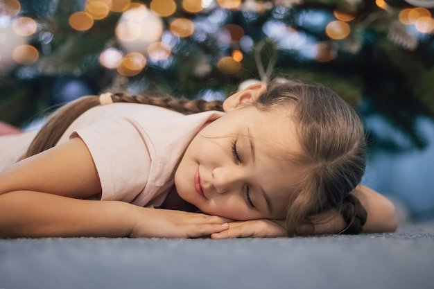 Close up shot of attractive small girl lies on floor, sleeps near New Year or Christmas tree, has pleasant dreams in evening. Children, winter holidays, rest concept