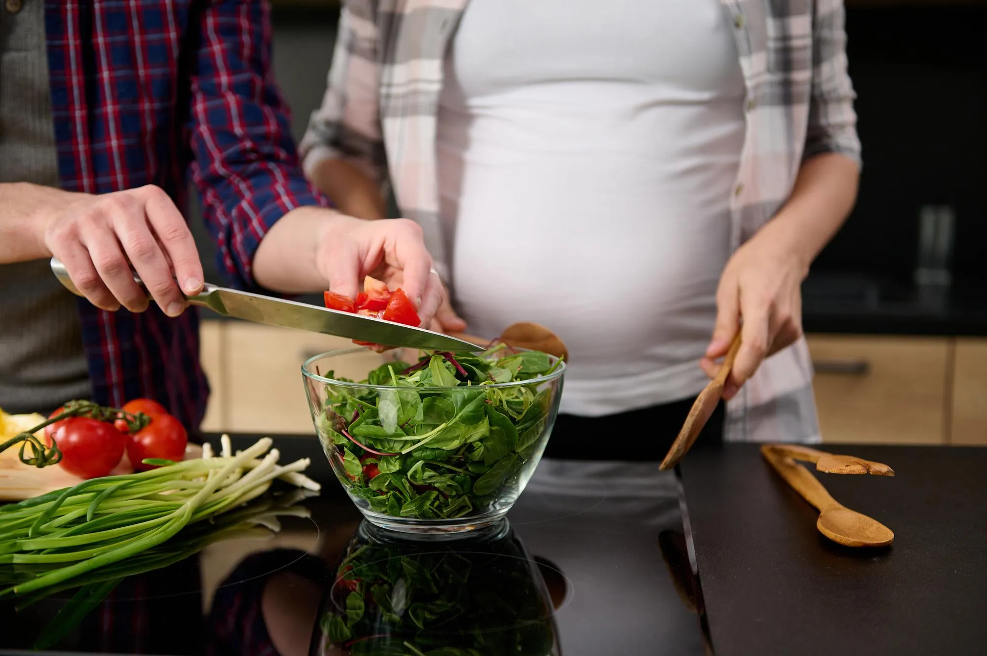 Focus on male hands holding a kitchen knife and puting sliced tomatoes into a bowl with lettuce leaves and greens on the background of his pregnant wife standing by a kitchen island.