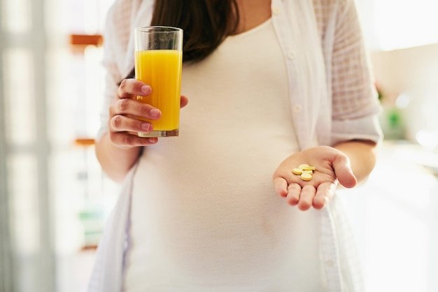 Hands pregnant woman and pills with juice for health supplements and start morning in home Person mother and orange drink for nutrition diet and pharma drugs for wellness benefits and change