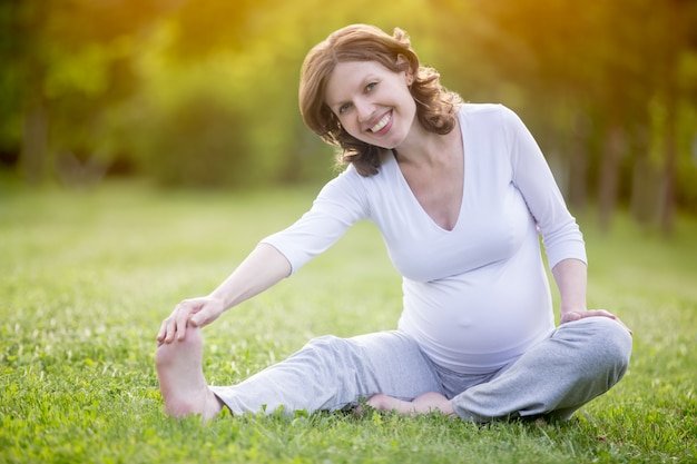 Happy pregnant woman doing stretching exercises on grass in park