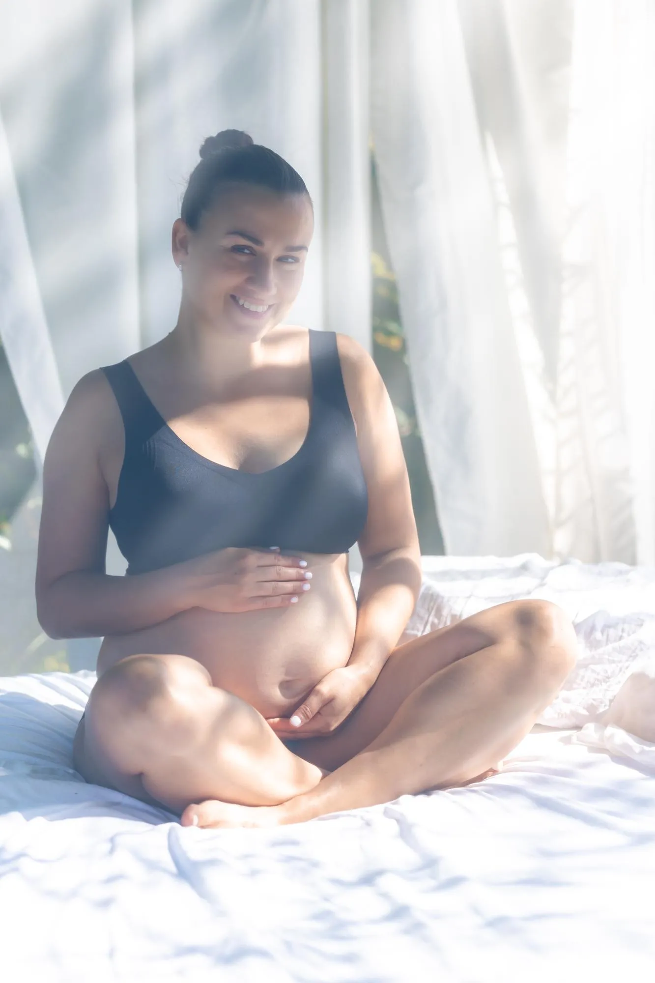 Happy pregnant woman touching her belly while sitting on a white bed