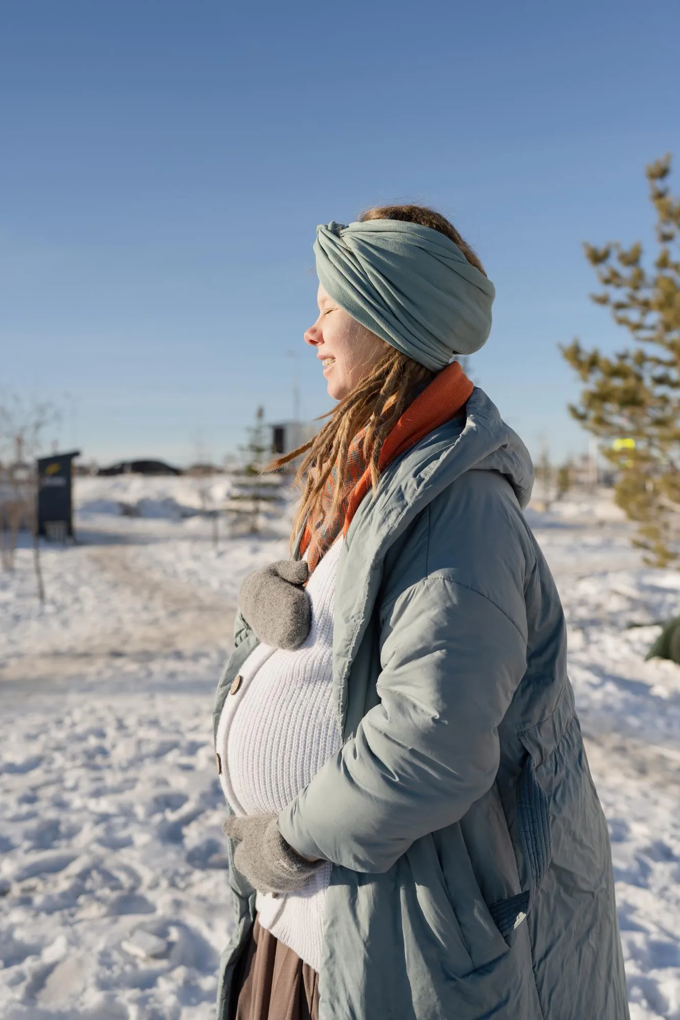 A happy pregnant young woman with her eyes closed, hugging her stomach in the park in winter