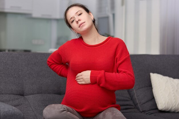 Photo of tired displeased pregnant woman feels backache, keeps hand on swollen belly, sits at couch with cushion, wears red loose sweater