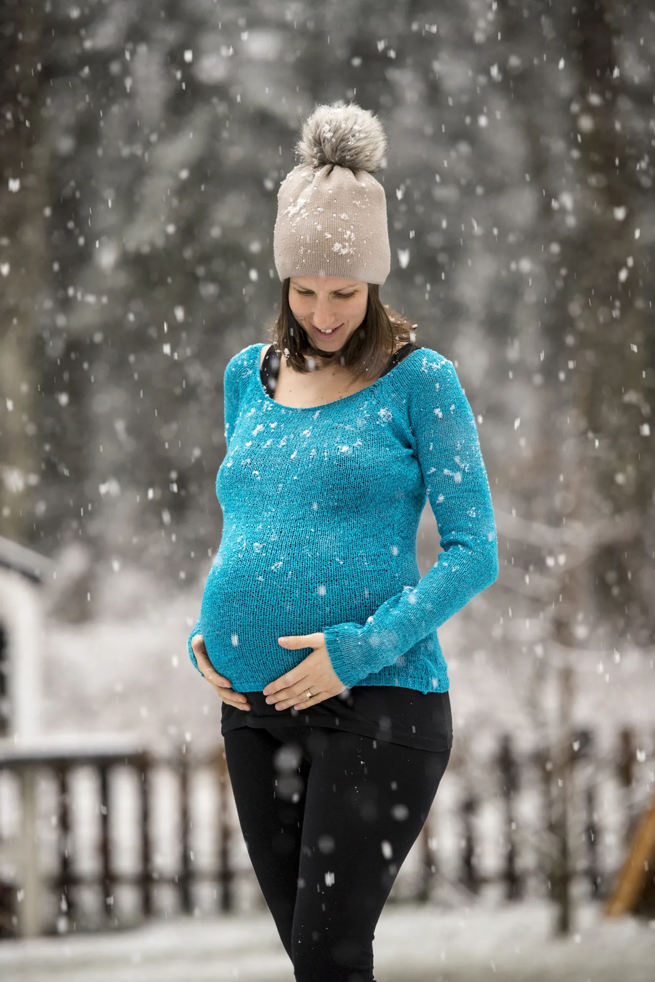 Pregnant woman holding her belly standing outside during snowfall wearing hat and blue sweater