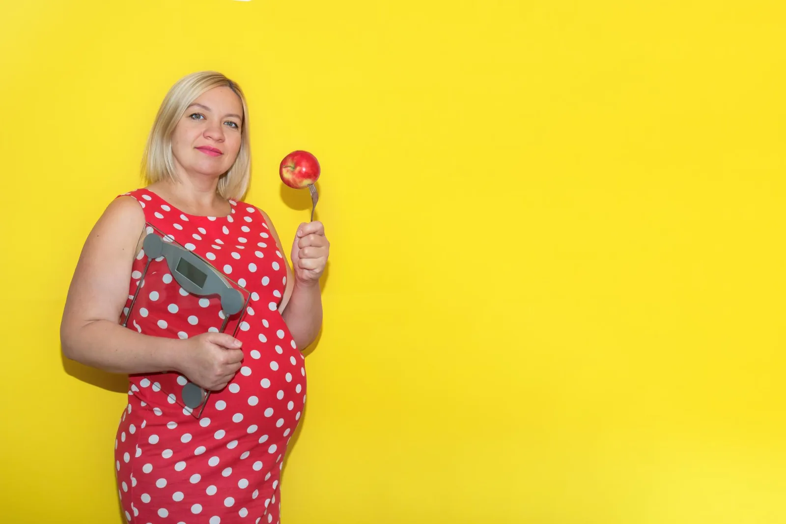 Pregnant woman in a red polka dot dress with scales and an apple on a fork on a yellow background with copy space