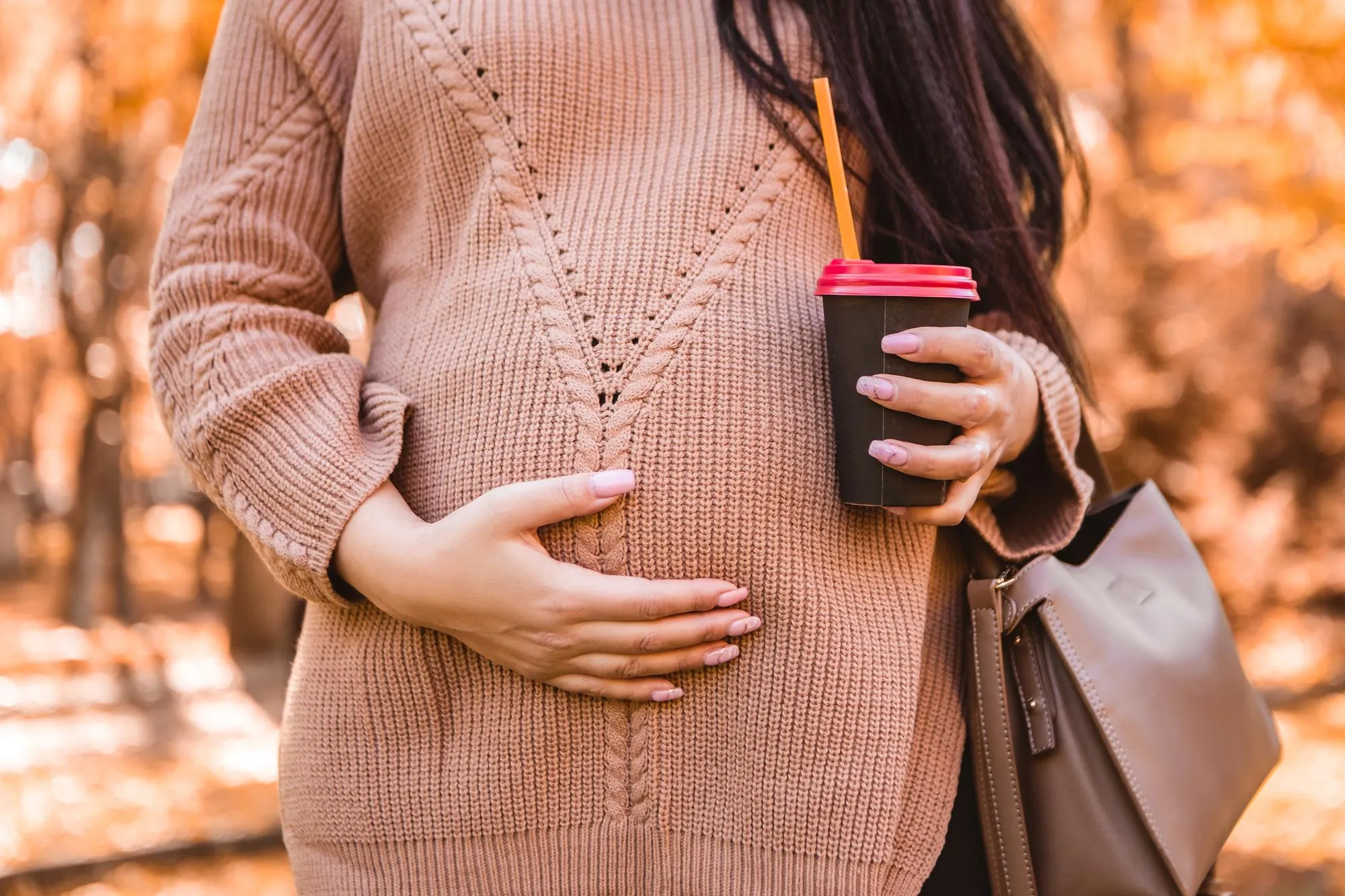 Pregnant woman standing in autumn city park forest, stroking her round belly with baby child inside
