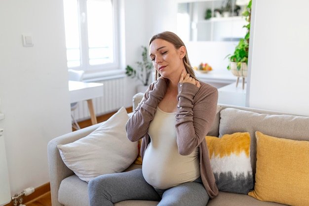 Pregnant woman suffer nrck and shoulder pain. pregnant woman holding her injured neck while touching her belly and sitting on couch