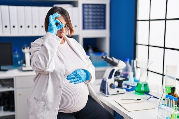Pregnant woman working at scientist laboratory doing ok gesture shocked with surprised face eye looking through fingers unbelieving expression