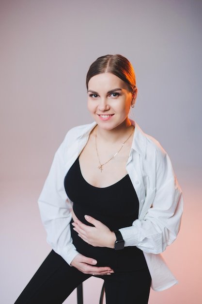 A stylish pregnant young woman in a white shirt and black pants sits on a chair and hugs her belly on a white background A pretty woman with professional makeup and casual clothes