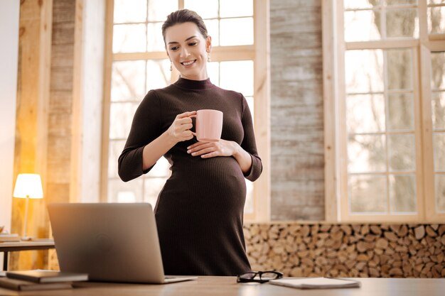 Tea time. Appealing pensive pregnant businesswoman holding cup while looking down and standing