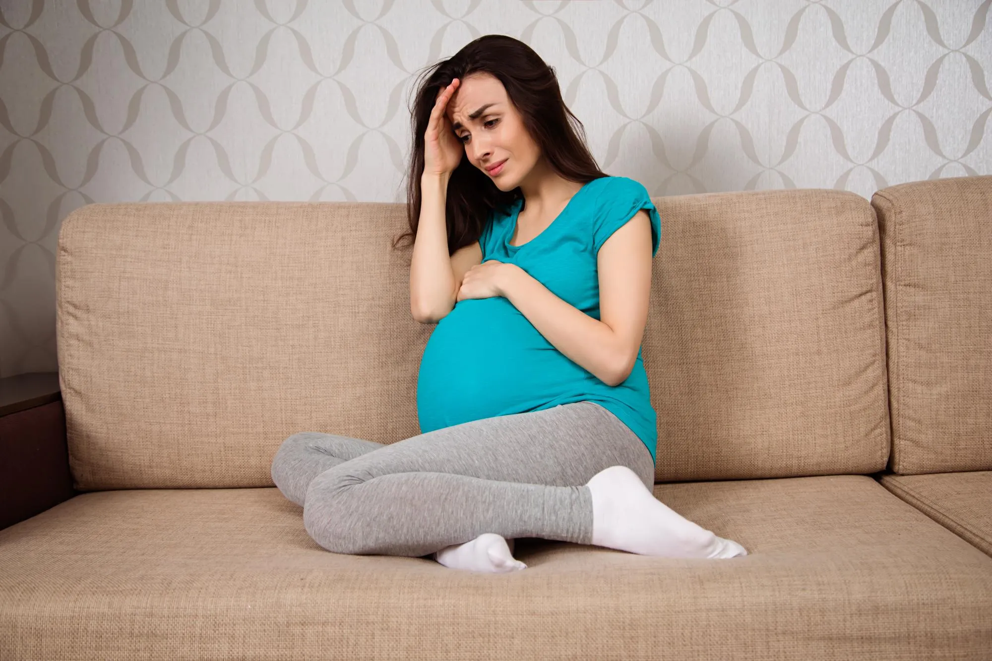 Upset young pregnant woman sitting on the couch at home and cries