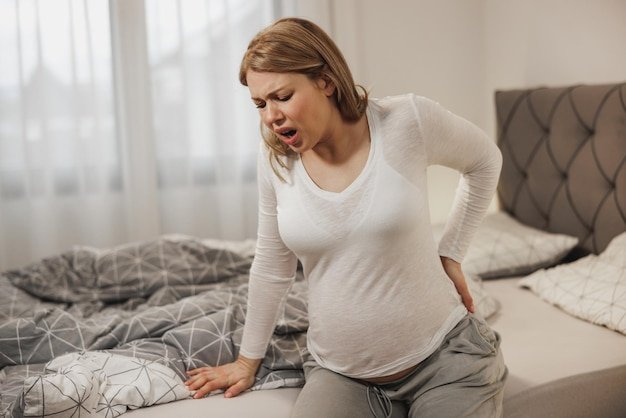 Worried pregnant woman holding her back in pain while sitting on a bed in bedroom.