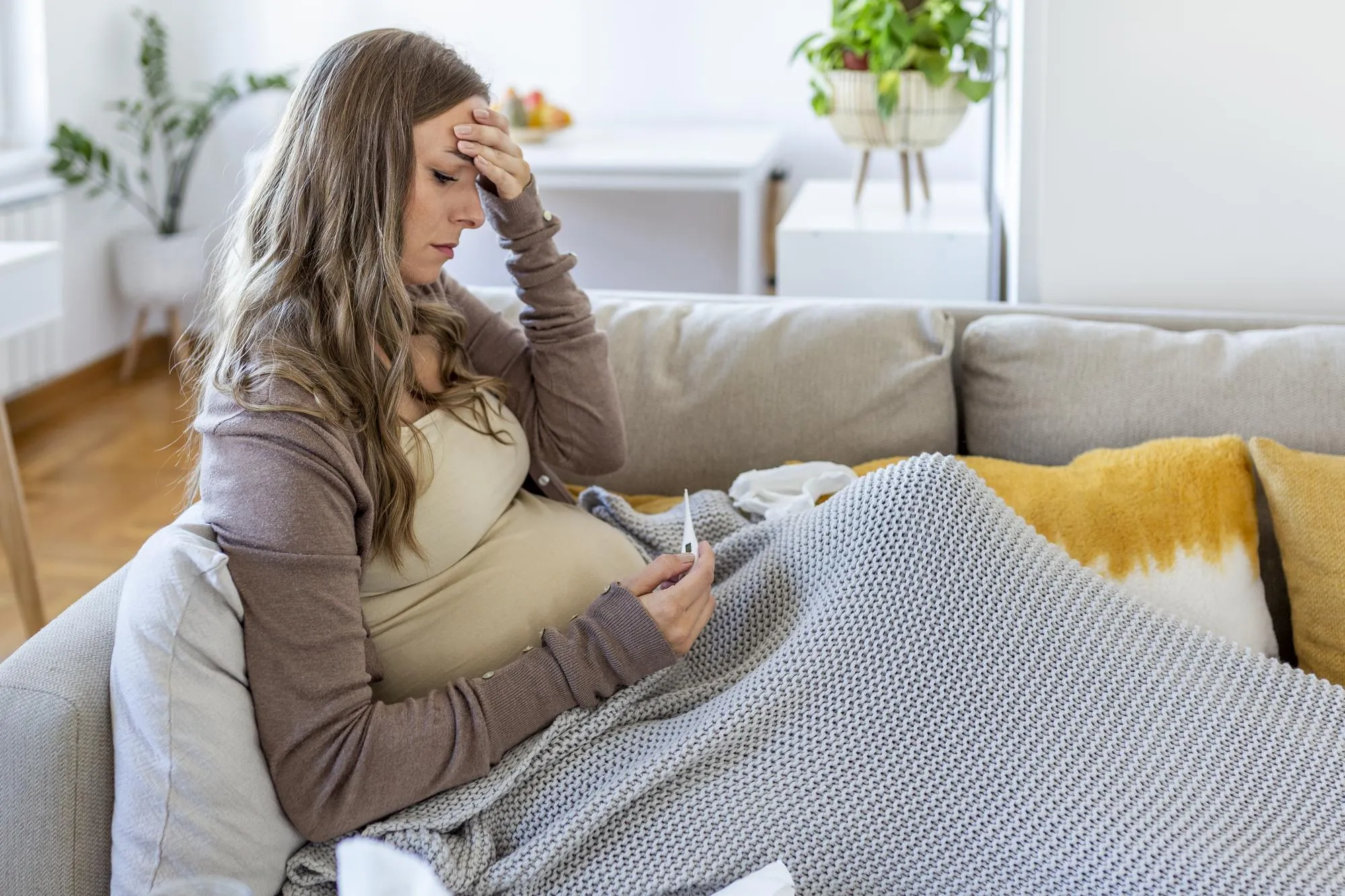 Young pregnant woman lying in bed measuring body temperature holding thermometer and face mask