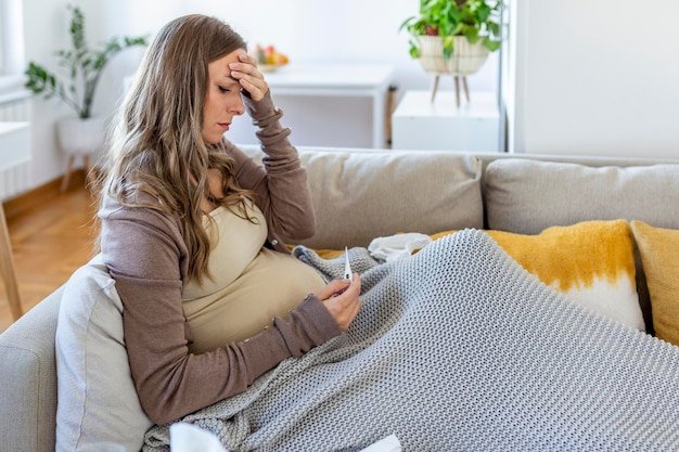 Young pregnant woman lying in bed measuring body temperature holding thermometer and face mask