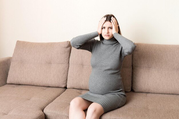 Young pregnant woman suffering from headache on sofa in home maternity healthcare concept