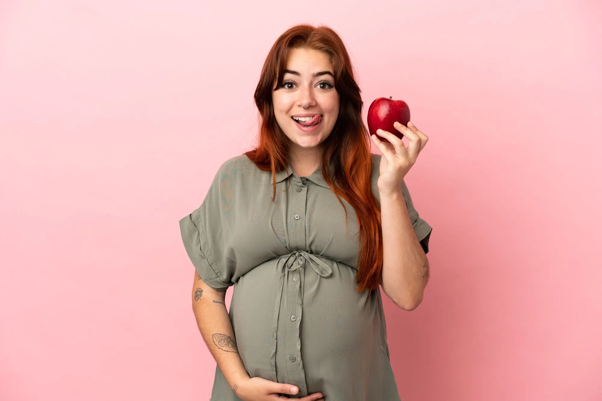 Young redhead caucasian woman isolated on pink background pregnant and holding an apple
