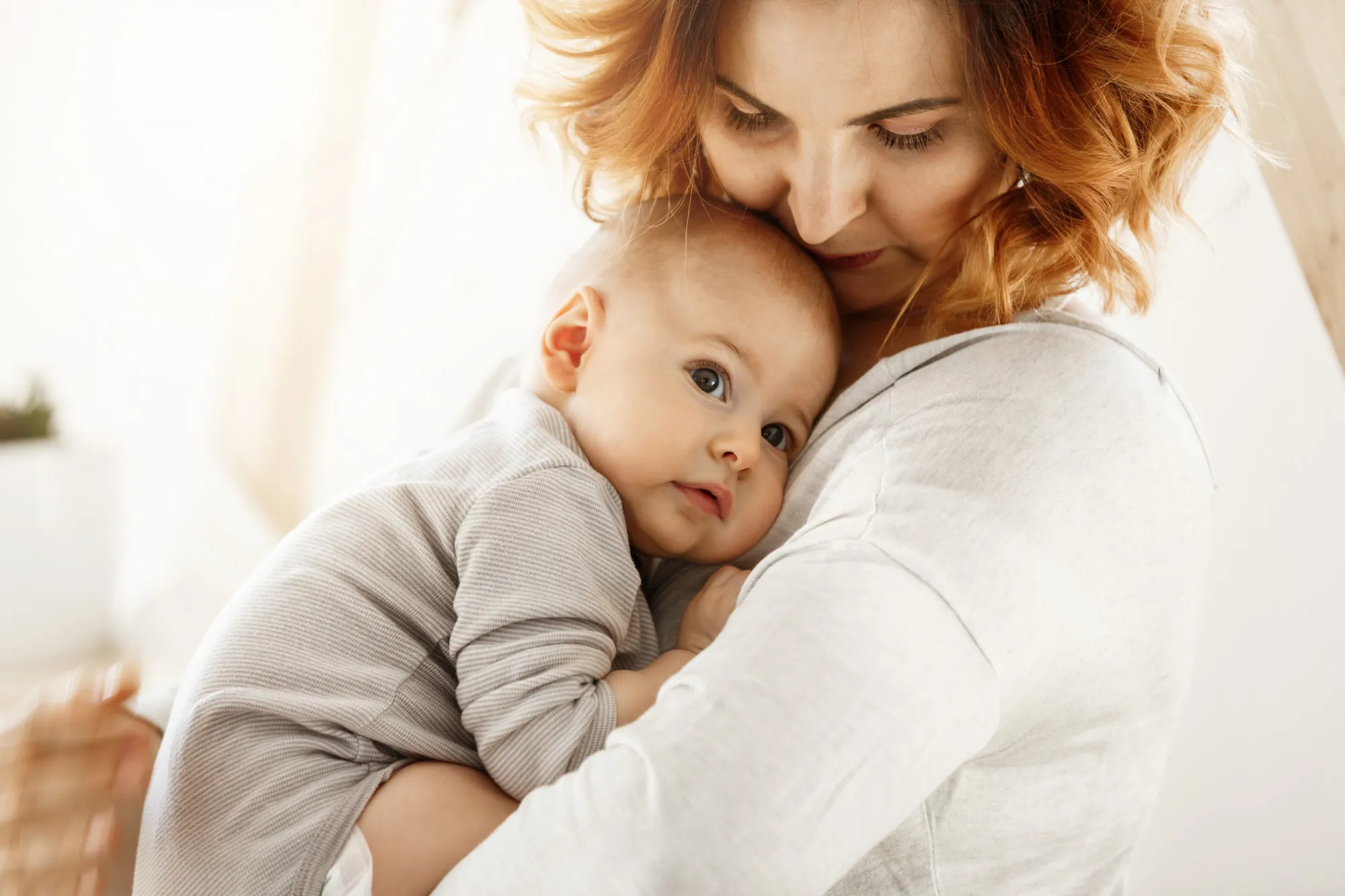 Beautiful young mother tenderly hugs her little cute child. Mom looking at her kid and smiling while baby looks aside and snuggling to mom chest. Happy family moments.