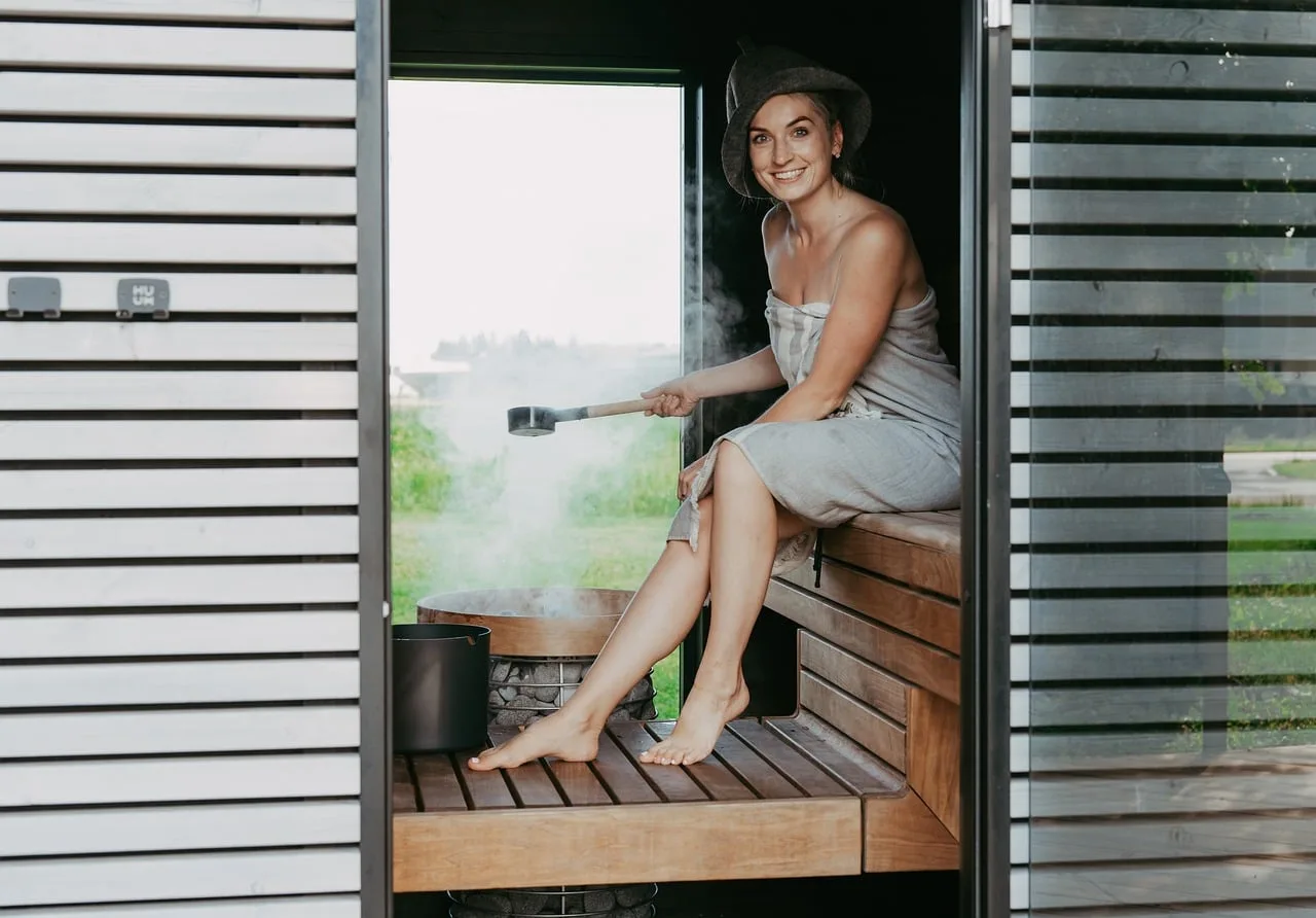 sauna, piec do sauny, kamienie do sauny, sauna parowa, grzejnik elektryczny, dom z sauną, projekt sauny, inspiracja sauną, nordycki design, bezpieczeństwo, barierka bezpieczeństwa, bezpieczeństwo w saunie, kobieta w saunie, siedzenie w saunie, śmiać się, osoba w saunie, ławka do sauny, nalewanie wody, grzejnik do ula, mini grzejnik do ula, kapelusz do sauny