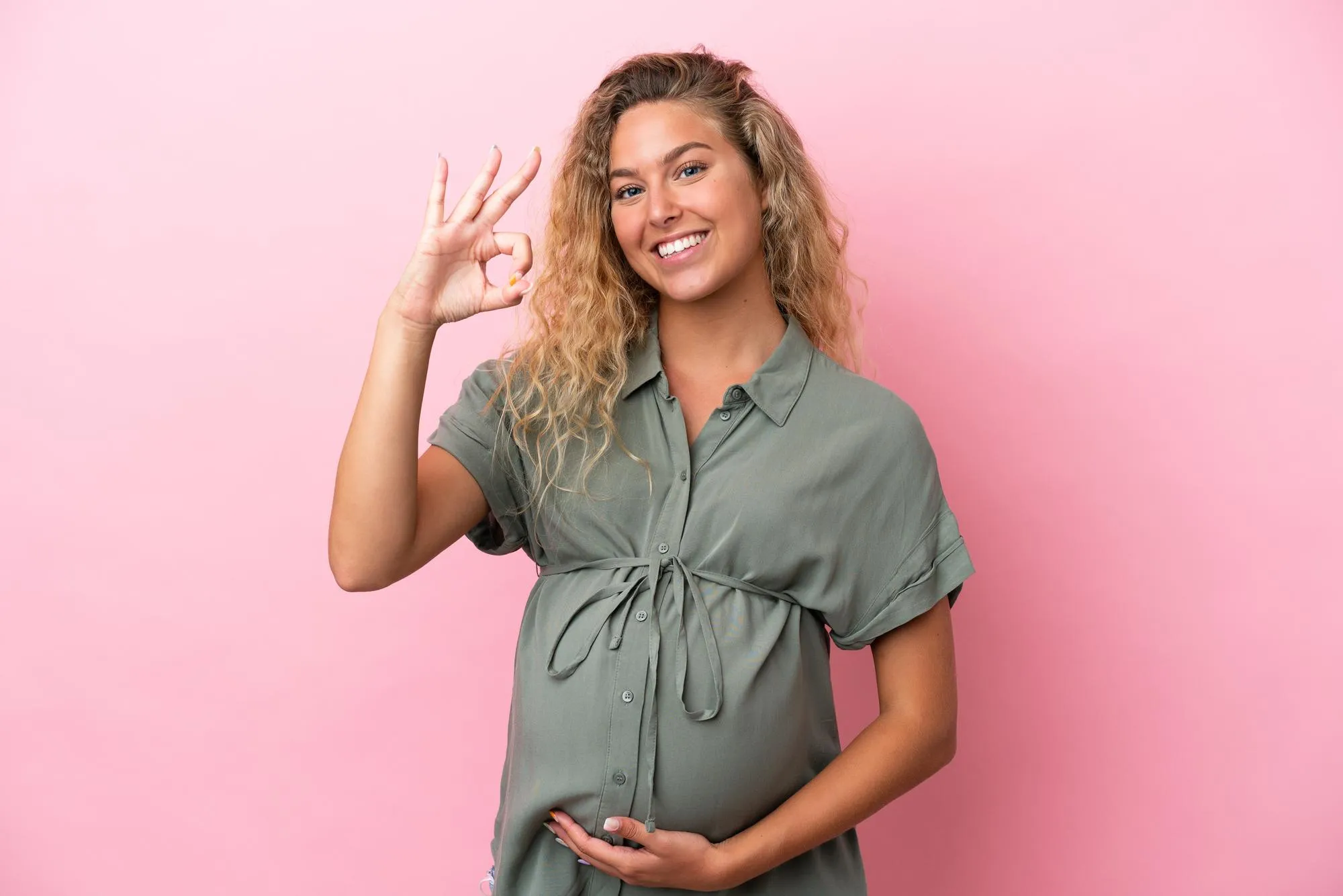 Girl with curly hair isolated on pink background pregnant and doing OK sign