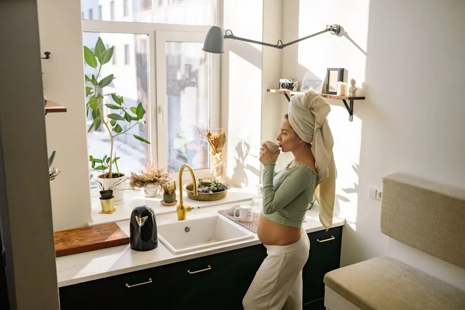 Pregnant woman enjoying a peaceful moment with tea by the window in a sunlit kitchen.
