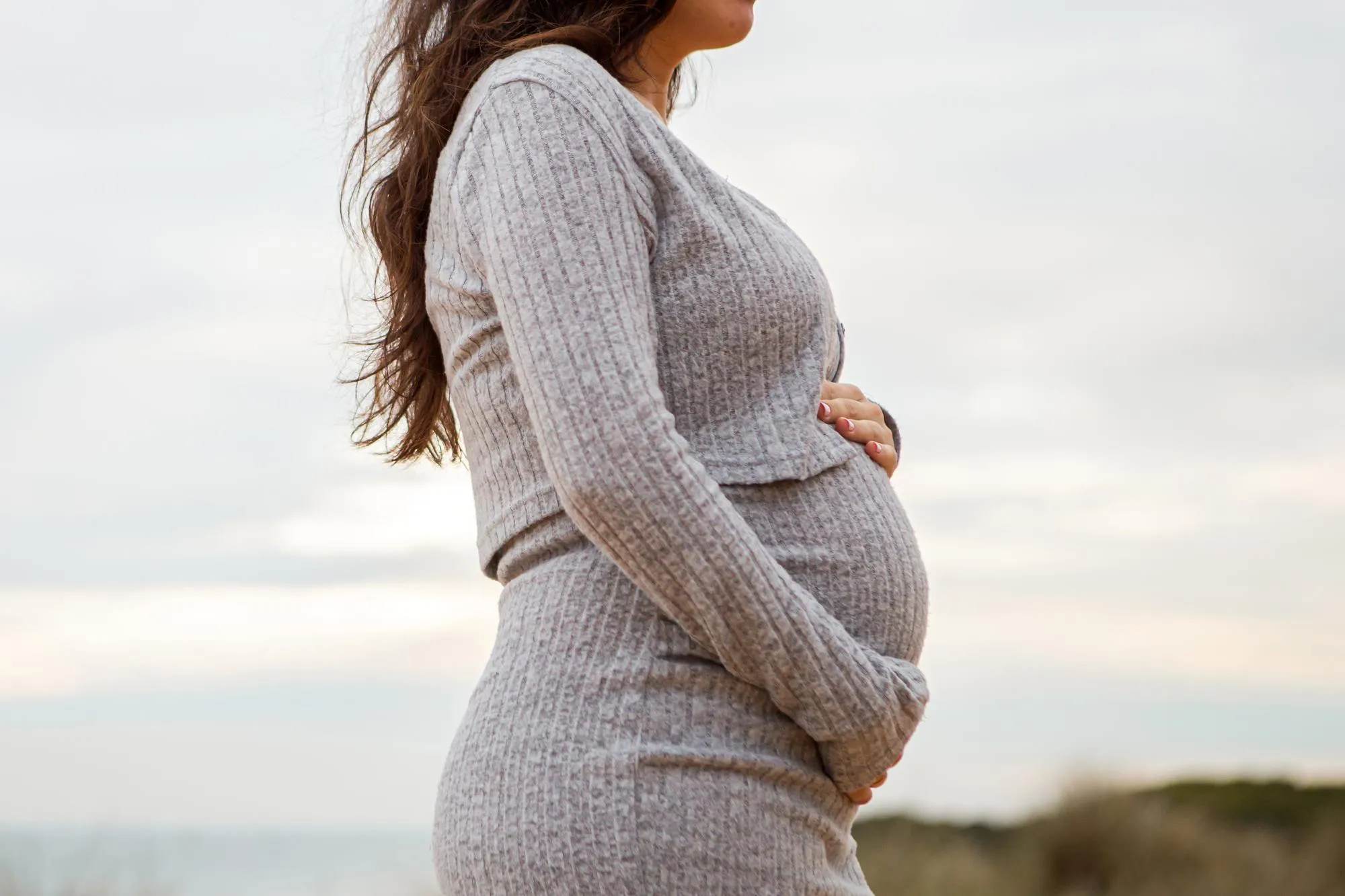 a pregnant woman stands on a beach with the sun setting behind her
