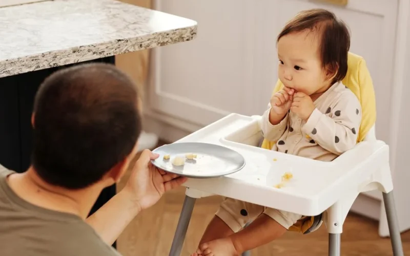 Adorable baby in a high chair eating while a parent offers food, indoors.