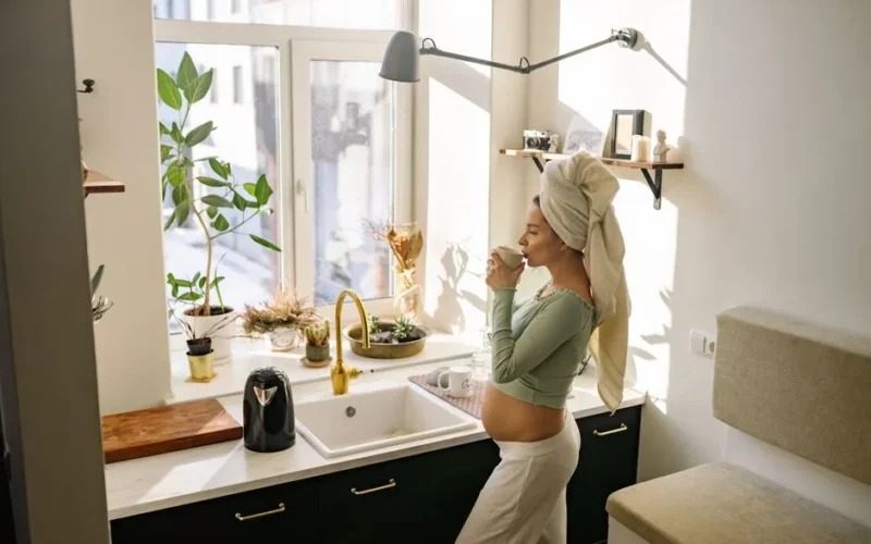 Pregnant woman enjoying a peaceful moment with tea by the window in a sunlit kitchen.