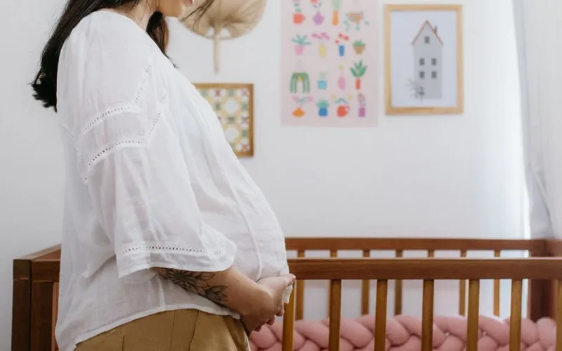 Expectant mother standing beside a wooden crib in a beautifully decorated nursery room.