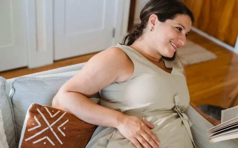 Young pregnant female in beige dress smiling and looking away while reading book on comfortable sofa