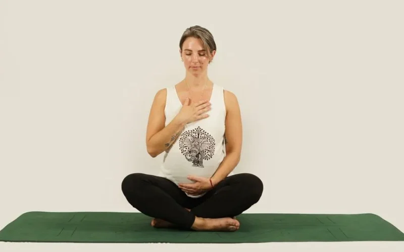Pregnant woman sitting cross-legged on a green yoga mat, meditating with a calm expression indoors.