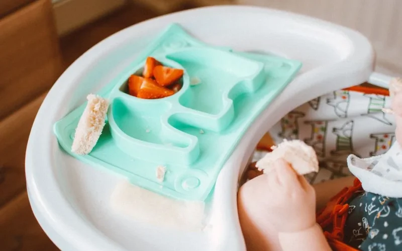 Adorable baby eating bread and carrots in a high chair with colorful tray.