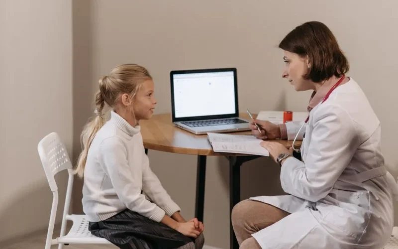 Doctor in lab coat consulting a young girl in a clinic with a laptop on the table.