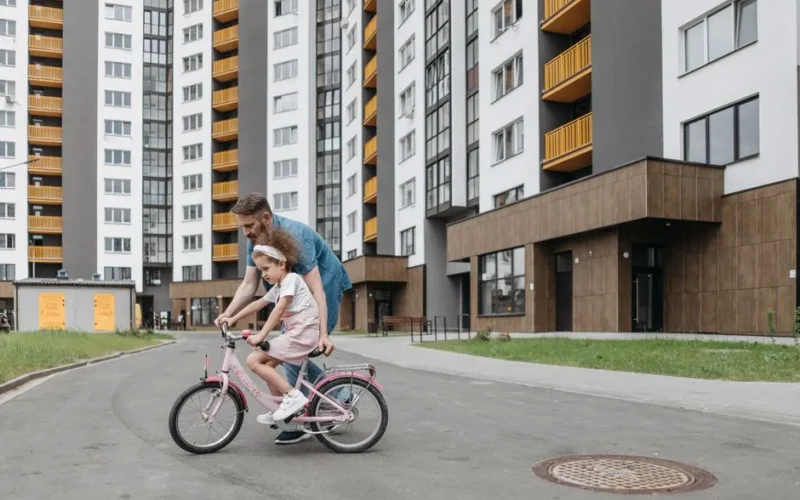 Father guiding daughter on a bike ride outside modern apartments. Bonding and teaching moment.