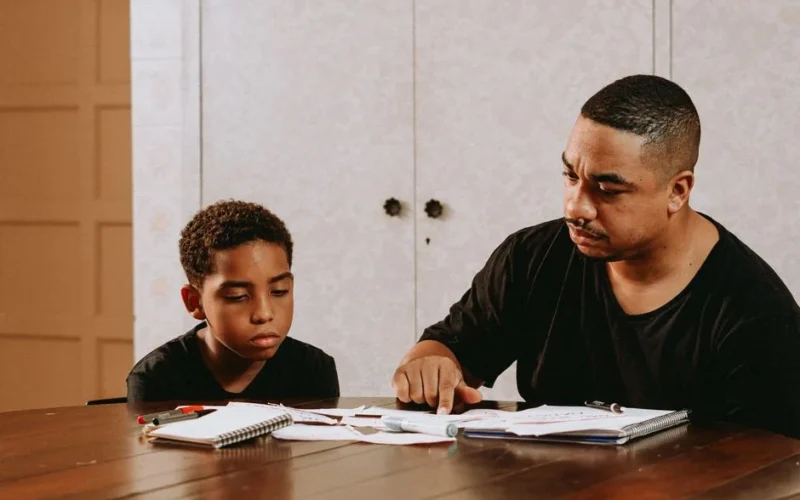 A father helps his son with homework at a dining table, promoting learning and bonding.