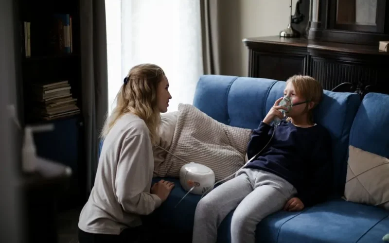 A mother assists her sick child using a nebulizer at home, providing comfort and care.