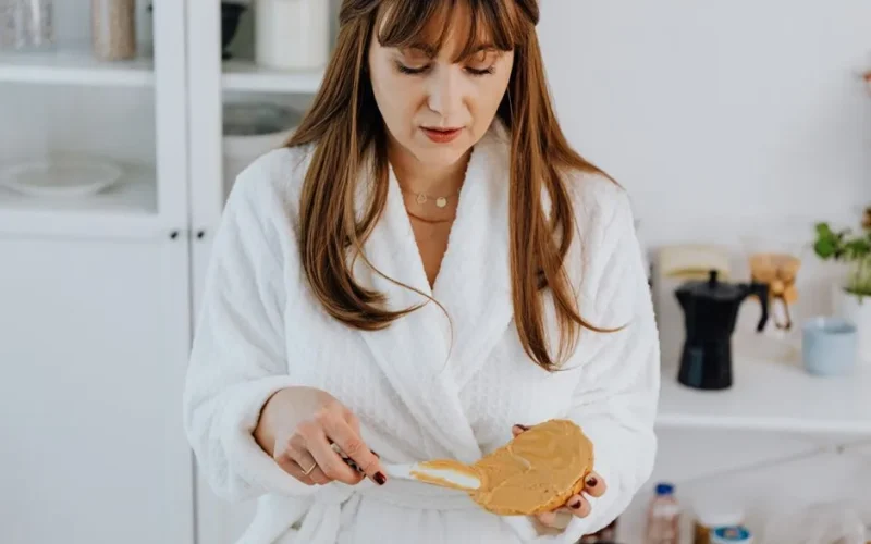 Woman in bathrobe spreading peanut butter on toast, indoors kitchen scene.