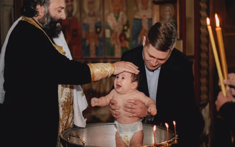 A priest performs a traditional baptism, blessing a baby in a church setting.