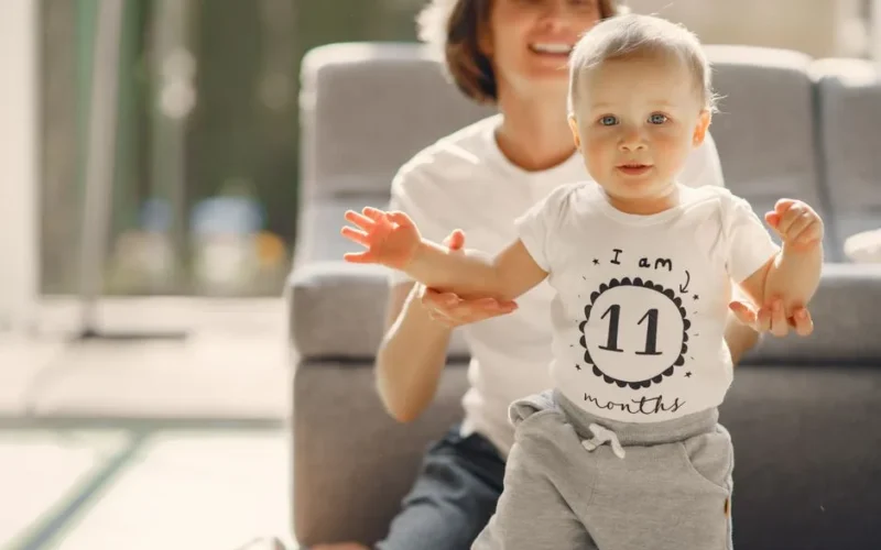 Adorable infant boy looking at camera while walking with support of loving mother in well lit living room