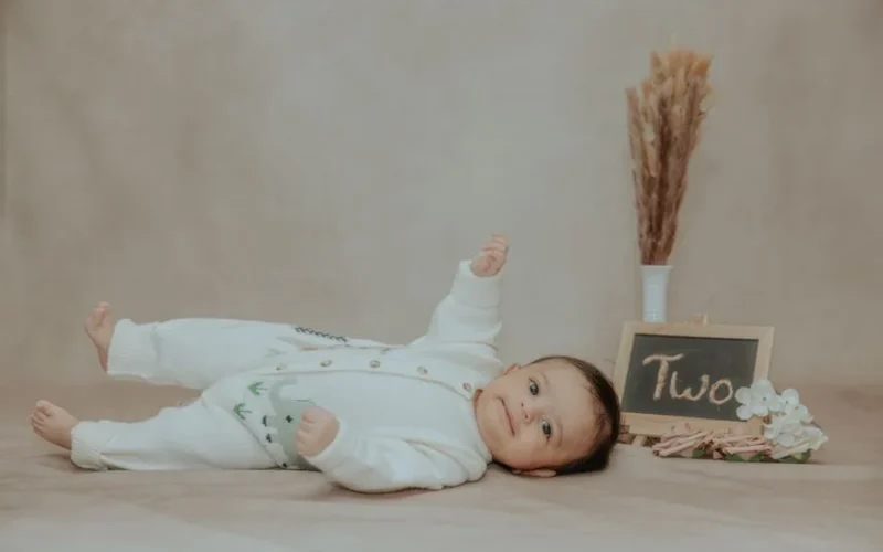 A cute two-month-old baby lies on a soft surface with a sign reading 'Two', surrounded by dried flowers.