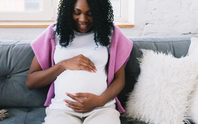 Cheerful pregnant African American female touching tummy gently while sitting on comfortable sofa with pillows in light room at home
