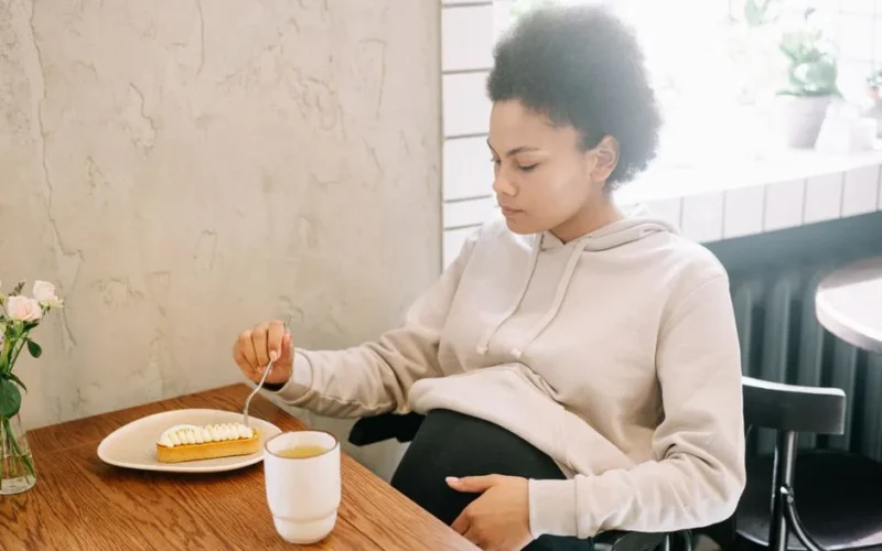 Serene pregnant woman in a cozy setting enjoying dessert and a hot drink.