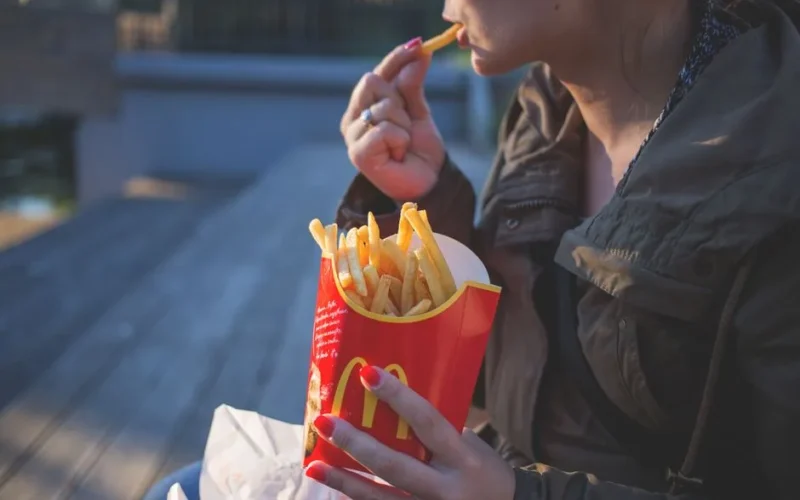 Hand holding french fry, bowl of fries, dipping sauce on table.