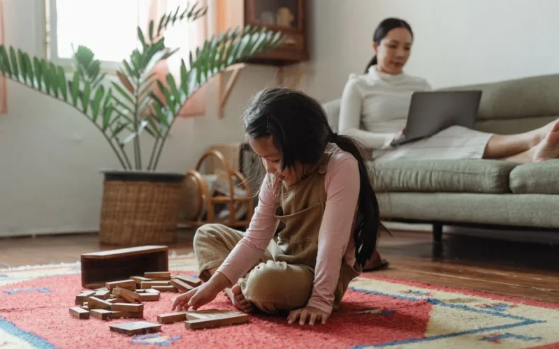 Content little Asian girl in casual wear sitting on carpet on floor and playing tower game while focused mother working on netbook on sofa near houseplant