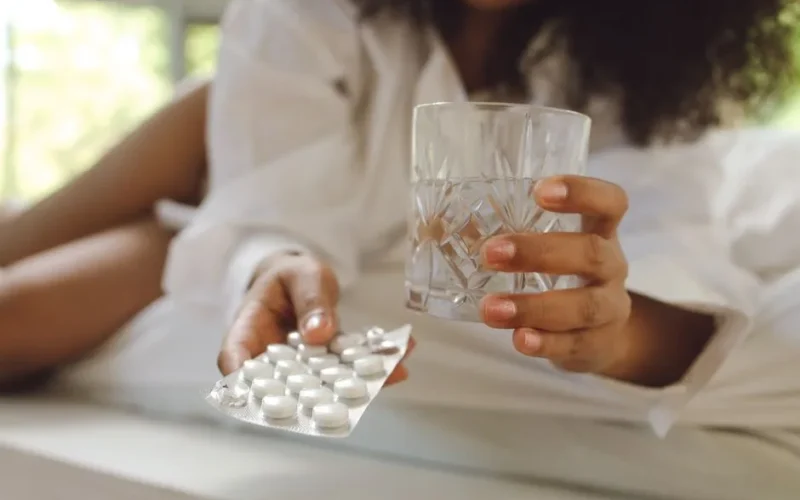 A woman in white long sleeves holds medication and a glass of water. Indoor lifestyle scene.