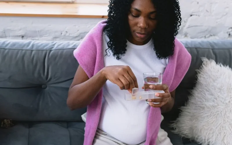 Serious pregnant African American female with glass of water taking prenatal vitamins while sitting on comfortable sofa in living room