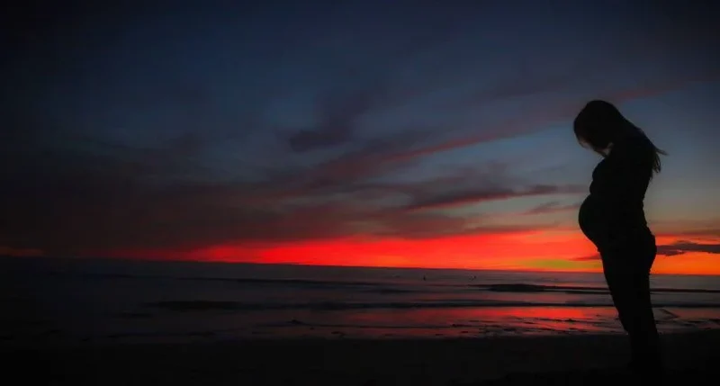 Silhouette of a pregnant woman by the beach during a vibrant sunset, capturing serenity and expectation.