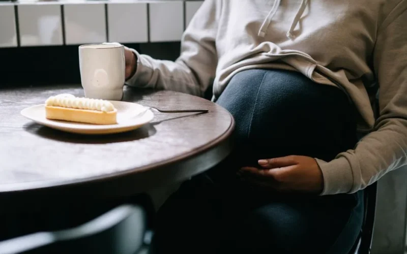 A close-up of a pregnant woman sipping coffee and enjoying a pastry indoors.